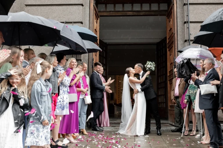 Jaunavedžiai bučiuojasi prie bažnyčios durų, svečiai su skėčiais aplinkui – vestuvių akimirka, Birute Bras Photography