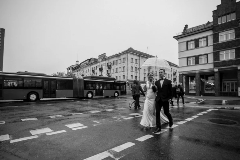 Jaunavedžiai kerta gatvę lietuje su skėčiu – juodai balta miesto vestuvių nuotrauka, Birute Bras Photography